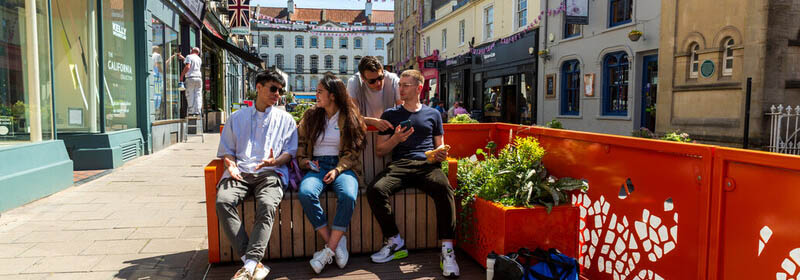 Four students sat together on a bench on a colourful, pedestrianised street in Clifton Village. 